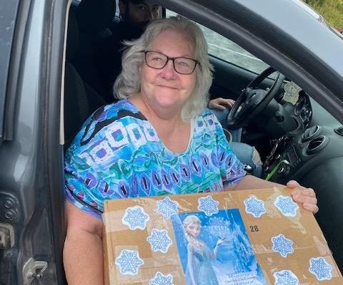 WILS Consumer, Ellen sits in her vehicle in a blue shirt, smiling.  She is holding a large cardboard box of produce, decorate with "Frozen" movie pictures.