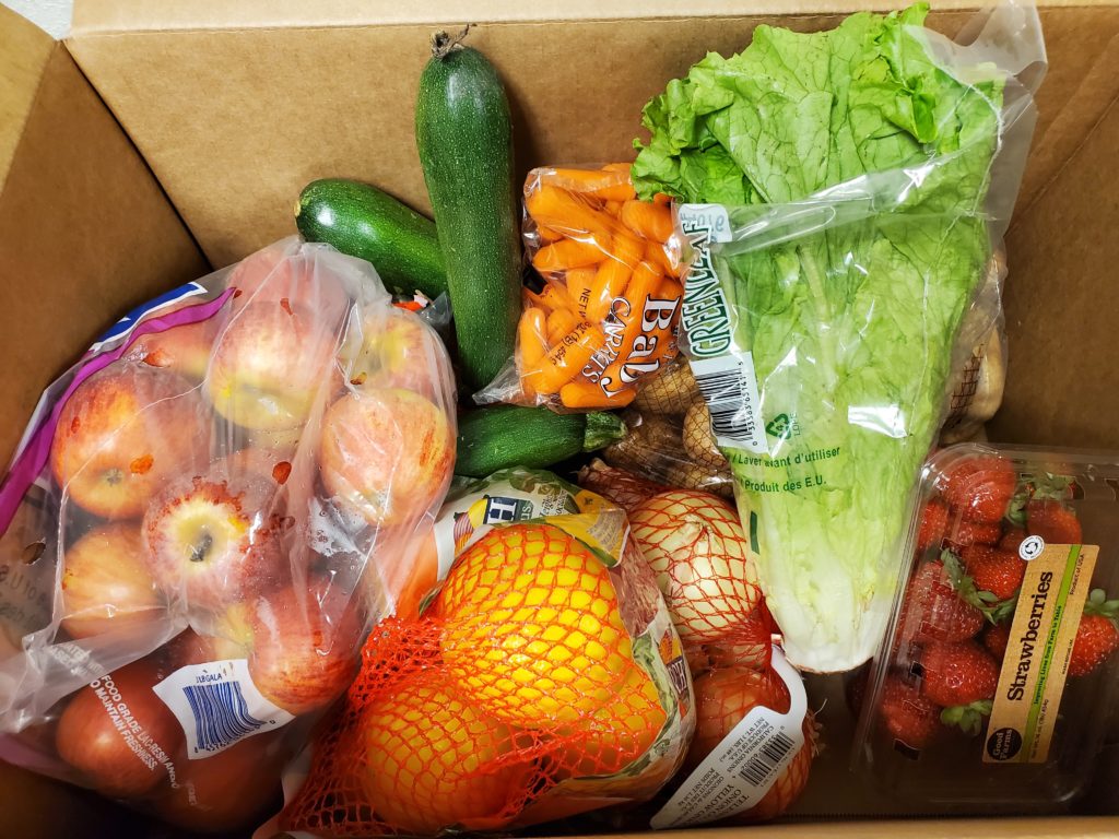 Cardboard box filled with fresh produce: apples, cucumbers, carrots, romaine lettuce, oranges, and strawberries.