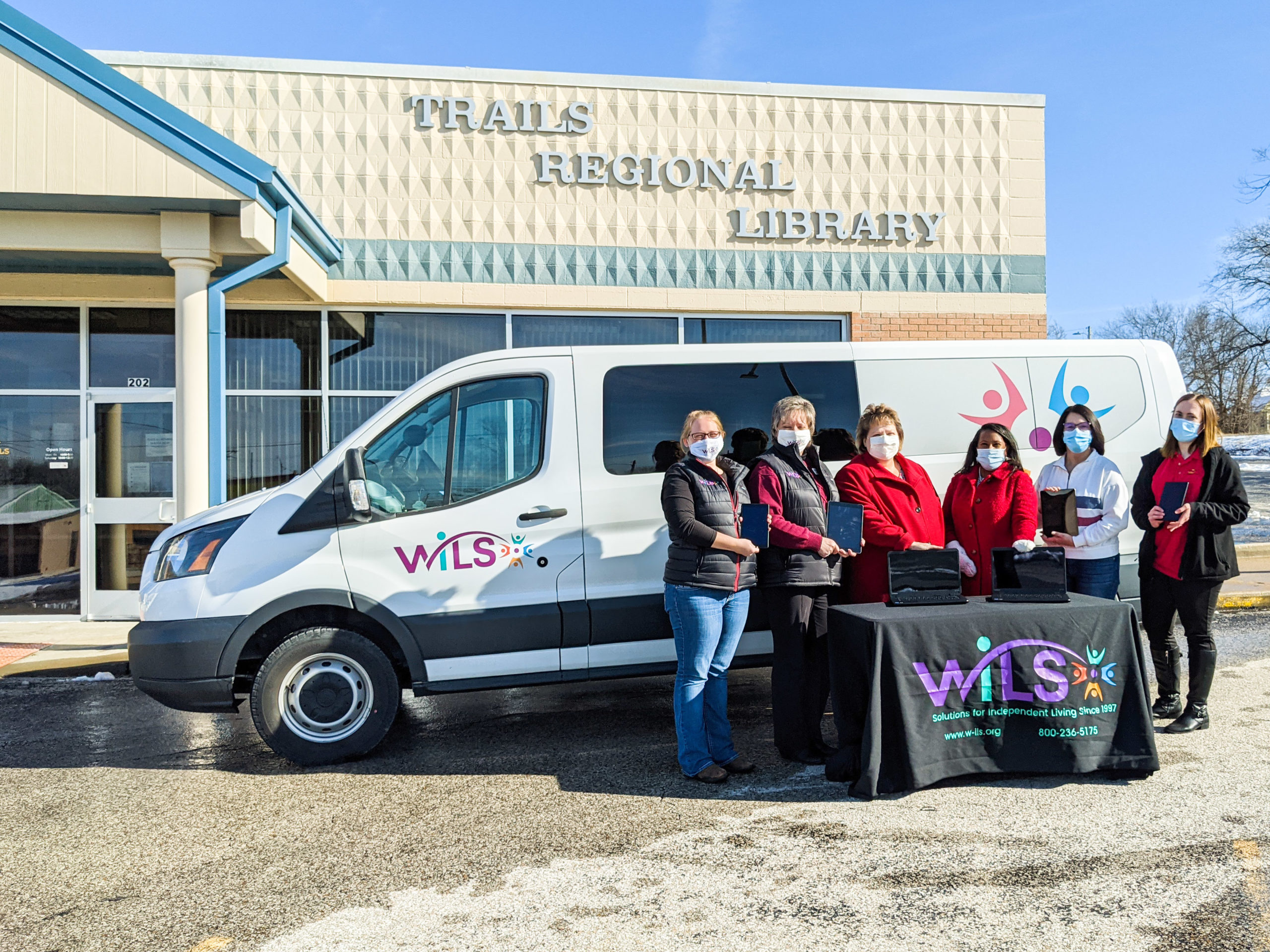 Ladies from Trails Regional and WILS stand behind a WILS table, in front of a WILS van that is parked in front of the Trails Regional Library in Knob Noster