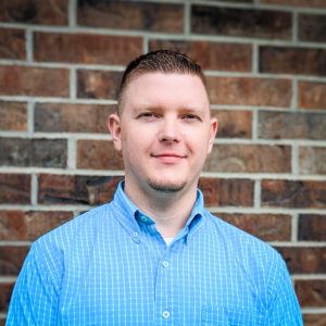 Photo of Bradley Callihan. He has brown hair and is standing against a brick wall and has on a blue collared shirt.