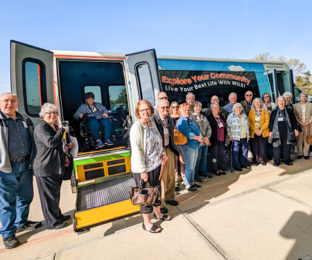 Group of people of various ages and abilities standing in front of WILS bus which reads, " Explore your community. Live your best life with WILS!"
