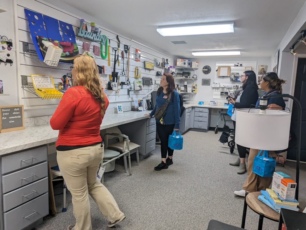 4 ladies look around in the AT Demo Center at WILS.  Slat boards are filled with kitchen, dressing aid, craft, office, communication, and visual aids.