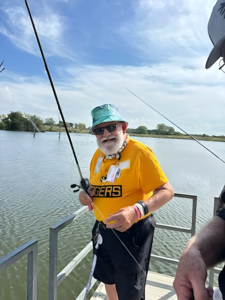 White male senior wearing a yellow tshirt, dark sunglasses, and turquoise bucket hat smiles while holding a fishing pole on a lake dock.