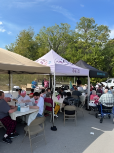 Large crowd of people sit at around tables under tents in a parking lot while eating a picnic lunch.