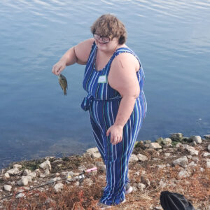 White female wearing a navy and white striped romper smiles while holding a fish on the lake shore.