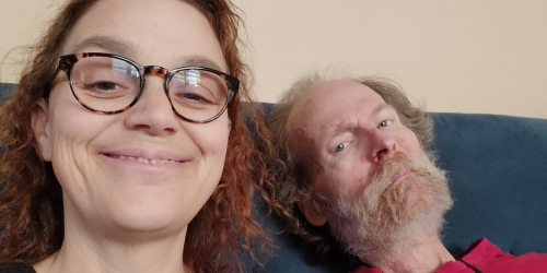 Daughter and father sit next to each other on couch. Both are white. Daughter has red wavy hair and glasses. Father has gray hair and a beard and is wearing a red shirt.
