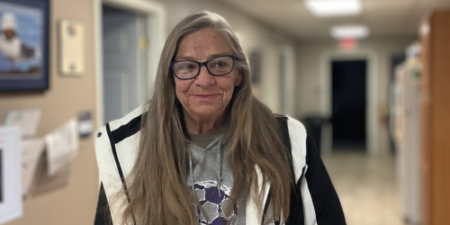 White woman with long, straight, dark blond hair wearing glasses, a gray hoodie, and a black and white winter coat is standing in a hallway smiling.