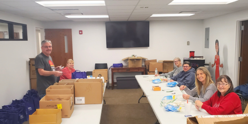 WILS volunteers are sitting and standing around tables filled with boxes and bags of community care supplies such as personal care and cleaning items.