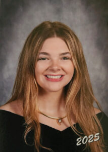 Photo is a headshot taken in front of a gray background. White female teen with blond hair wearing a black shirt and a necklace smiles into the camera.
