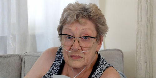 74 year old woman wearing a black and white tank top looks at the camera. She has short gray hair, is wearing glasses, and using oxygen.