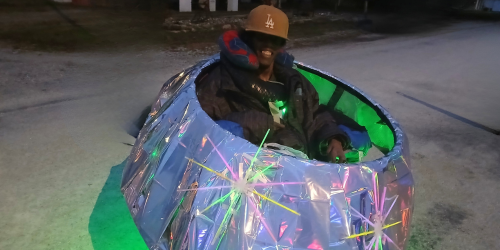 Man using a whellchair wearing a silver flying saucer costume, black coat, and tan LA hat.