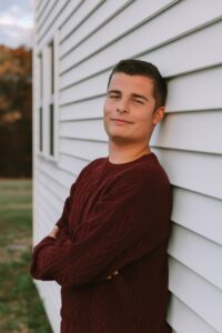A male high school senior with brown hair wearing a red-brown shirt leans up against a white-sided building and smiles at the camera.